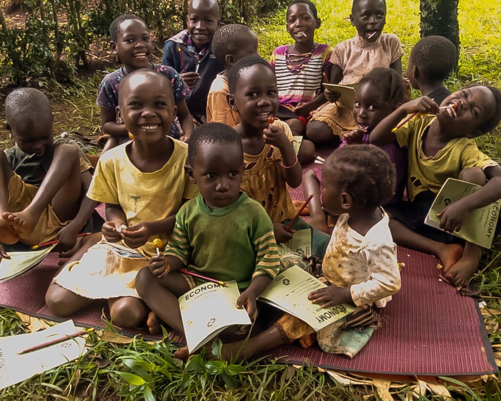 Children sitting together on a mat outdoors, reading and writing in exercise books.